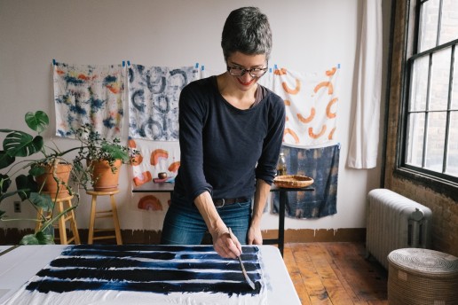 A textile artist hand paints blue stripes across a white silk scarf