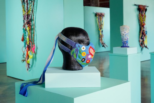 In the foreground is a beaded mask with a colorful butterfly motif placed on a black mannequin head form. In the background on three different walls that are turquoise green hang large, colorful, knotted rope works. In the midground to the right is a beaded sculpture with blue and white circular motifs that rests on a turquoise green pedestal.