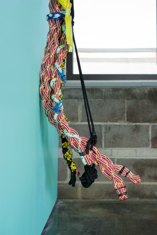 A side profile of one of Liz Miller's knotted rope sculptures hangs on a turquoise green wall on the left. The sculpture curves up and out from the wall at the base.