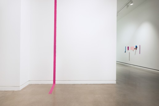 In the foreground to the left, a straight magenta line stretches from the top of the ceiling and extends onto the floor towards the viewer. On a back wall to the right, various paper sculptures in blue, magenta, and pink, are displayed on the wall.
