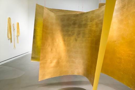 Three large square forms are suspended from a ceiling in a cluster in the middle of a room. They have yellow backs and gold foil fronts. On a white wall to the left, Rectangular gold foil forms drape and loop along the wall.