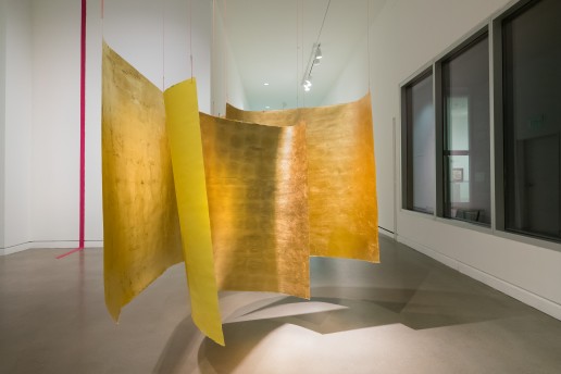 Three large square forms are suspended from a ceiling in a cluster in the middle of a room. They have yellow backs and gold foil fronts. On the back wall to the left, a magenta line stretches from the top of the ceiling and extends partially across the floor.